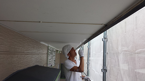 20130722_noki | 外壁塗装は神奈川県横浜市の塗装職人 Construction worker wearing a white hard hat and gloves applies sealant under scaffolding on an exterior building façade.