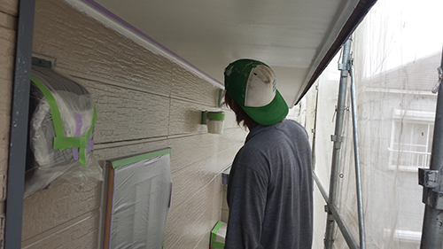 20130723_shitanuri_kazu | 外壁塗装は神奈川県横浜市の塗装職人 Person in a gray shirt and green cap painting the exterior wall of a house, with masking tape and plastic coverings on nearby surfaces under an overhang.