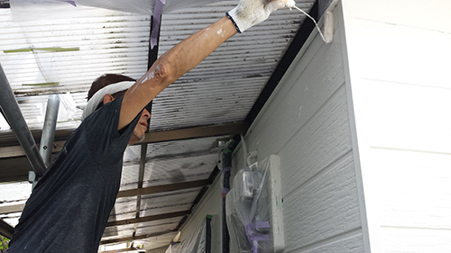 20130727_nakanuri2 | 外壁塗装は神奈川県横浜市の塗装職人 Worker in a black shirt and gloves applies paint to a corrugated metal ceiling from a scaffold or ladder area, wearing a mask for protection.