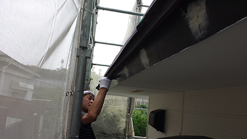730_hafu-shitanuri1 | 外壁塗装は神奈川県横浜市の塗装職人 Construction worker on scaffolding painting the underside of a building's overhang, wearing gloves and a hard hat.