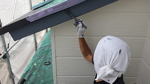 730_hafu-uwanuri1 | 外壁塗装は神奈川県横浜市の塗装職人 Worker in a hooded shirt spray-paints the exterior trim under the eave from scaffolding.