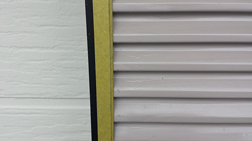 731_amado1 | 外壁塗装は神奈川県横浜市の塗装職人 Close-up of a pale green wall and a gray metal rolling shutter, divided by a vertical yellow edge.