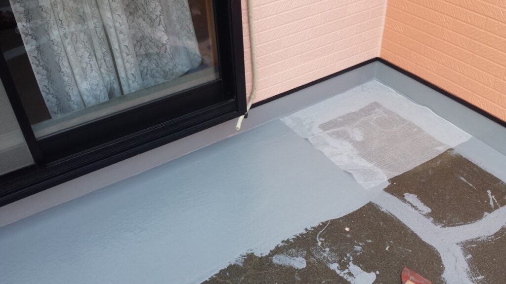METADATA-START | 外壁塗装は神奈川県横浜市の塗装職人 Balcony floor being coated with light gray waterproofing near a sliding glass door with lace curtains outside a pink wall.