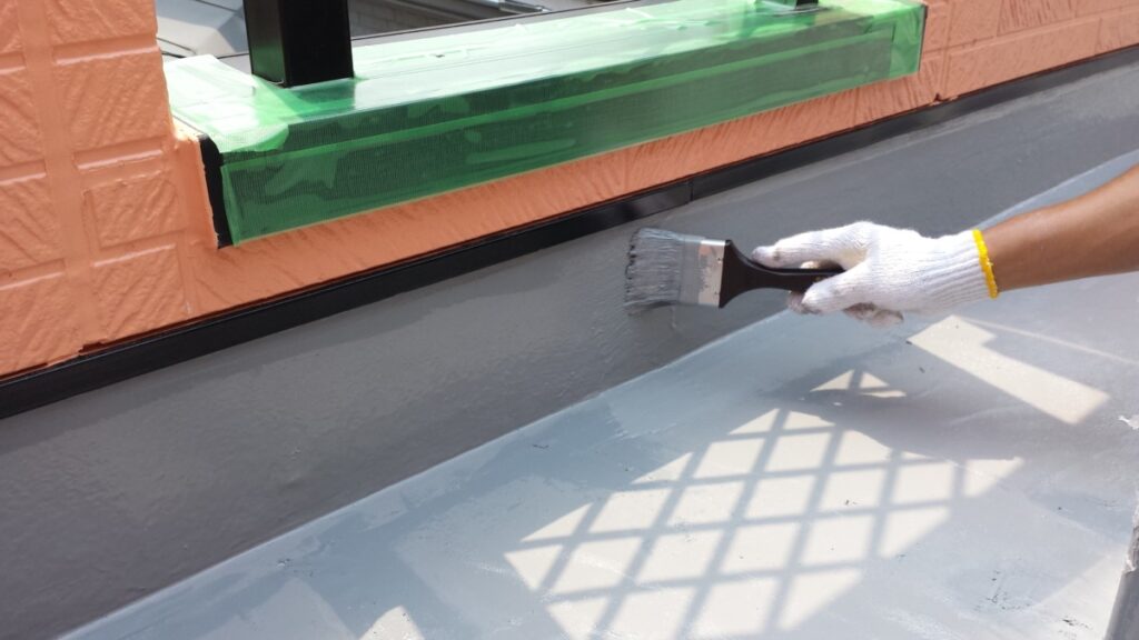 METADATA-START | 外壁塗装は神奈川県横浜市の塗装職人 Worker's gloved hand painting a roof edge with gray paint along an orange brick wall masked with green tape.
