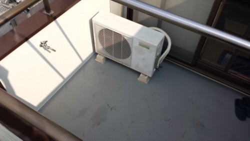 20130812-083626 | 外壁塗装は神奈川県横浜市の塗装職人 Outdoor air conditioning condenser unit mounted on a balcony floor, with railing and a pair of slippers in the corner.