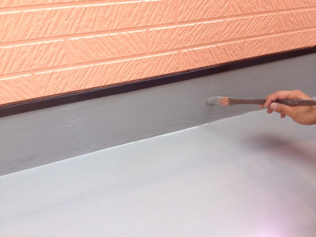813-image | 外壁塗装は神奈川県横浜市の塗装職人 Hand holding a paintbrush applying gray paint to a baseboard against a peach brick wall.