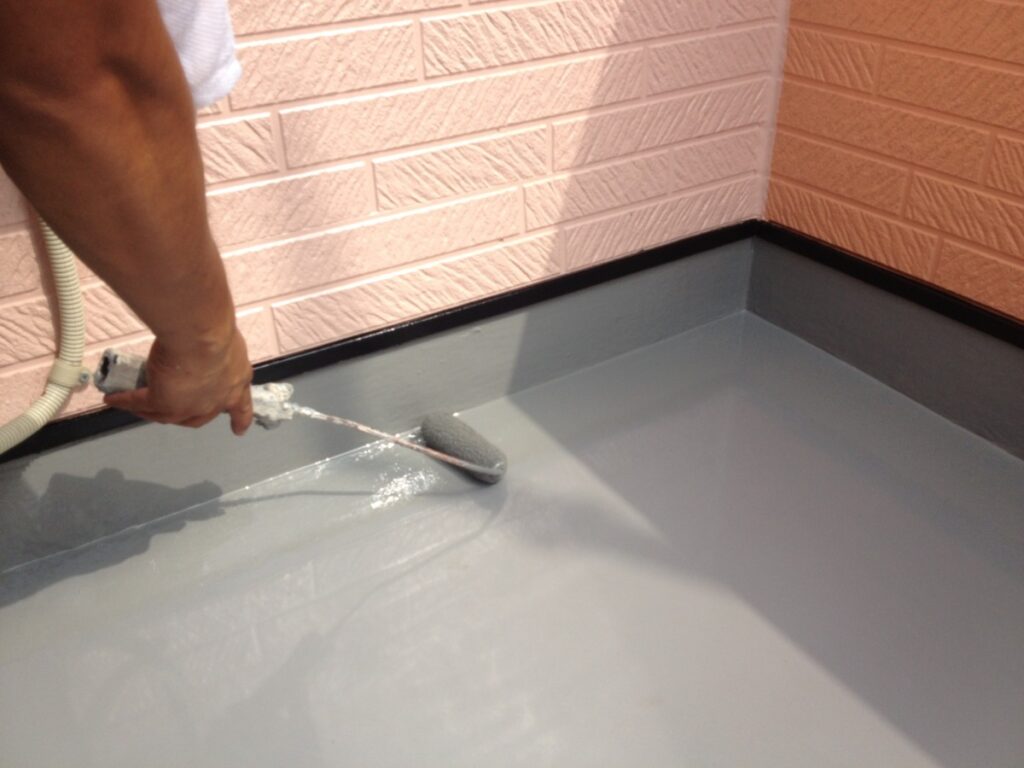 813-image_1 | 外壁塗装は神奈川県横浜市の塗装職人 Person using a paint roller to apply a gray coating on a floor in a corner surrounded by pink brick walls, near baseboard trim.