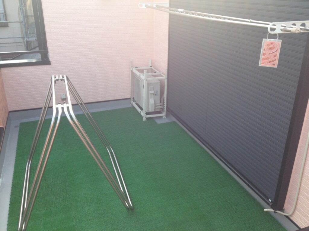 813-image_6 | 外壁塗装は神奈川県横浜市の塗装職人 Small balcony with green outdoor carpet, a metal triangular drying rack, a wall air conditioner unit, and a clothesline with a tag hanging from it.