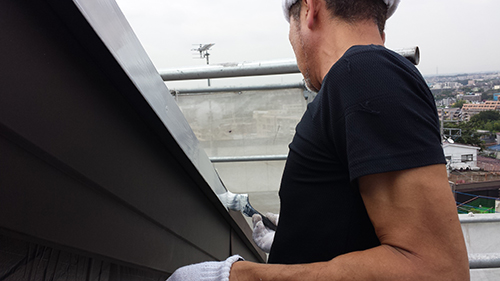 Man on a rooftop wearing a black T-shirt and gloves, working along the edge of a building with a cityscape in the background.