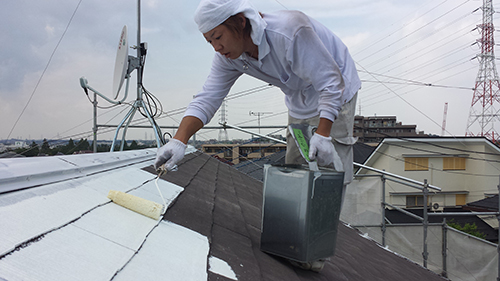 Roofer on a sloped residential roof applying white coating with a trowel, toolbox nearby.