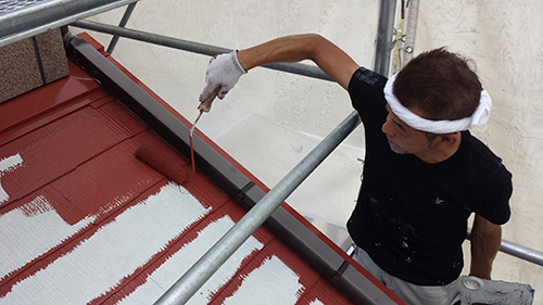Worker on scaffolding painting a red metal roof with white paint using a roller.