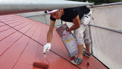 Worker on a sloped red roof applying paint with a roller, paint can in hand.