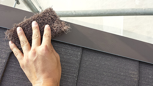 A hand presses a clump of brown insulation fibers onto a roof seam above dark shingles with scaffolding in the background.