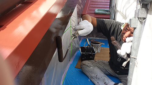 Tradesperson on a scaffold painting a metal beam with orange-red paint using a roller; blue tarp on the ground and a paint bucket nearby.