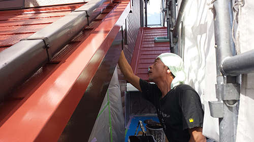 Construction worker on scaffolding applying sealant along the edge of a red metal roof.