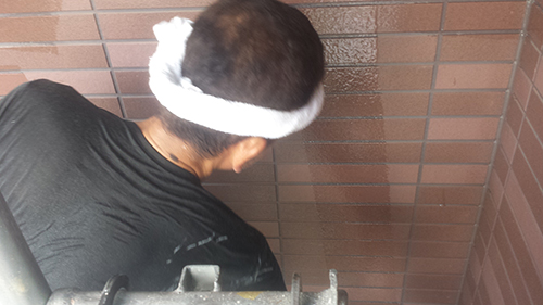 Back view of a person wearing a white cloth headband and a black shirt, standing against a brown brick wall in a narrow space.