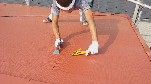 Worker wearing gloves trims red roofing material on a sloped roof with a utility knife and scraper nearby