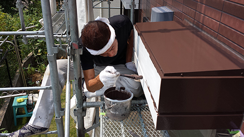 Construction worker on scaffolding painting a brown metal vent box on the exterior wall with a brush.