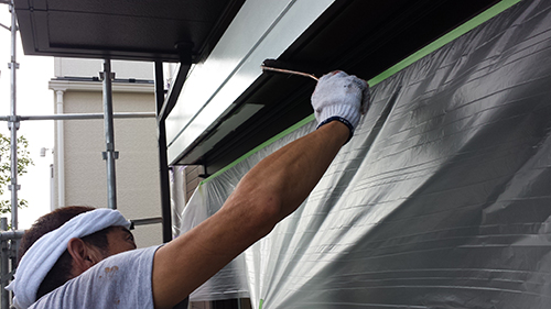 Worker with white headband painting a building exterior, protected by plastic sheeting.