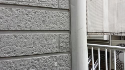 20130826-083603 | 外壁塗装は神奈川県横浜市の塗装職人 Close-up of a gray brick wall with a white vertical pipe and a metal railing in an urban building setting.