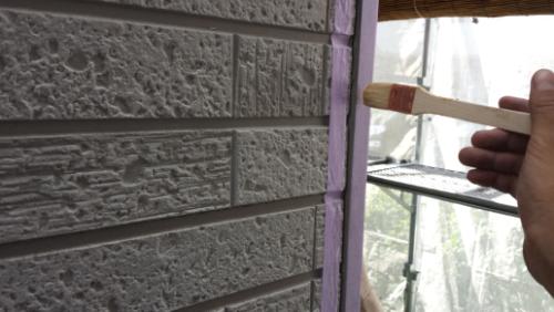 20130827-092430 | 外壁塗装は神奈川県横浜市の塗装職人 Person painting a joint along a brick exterior wall with a brush, purple masking tape nearby and scaffolding in the background.