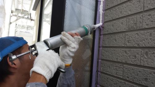 20130828-110645 | 外壁塗装は神奈川県横浜市の塗装職人 Construction worker in a blue cap and gloves applying sealant with a caulk gun along a window frame on a brick wall.
