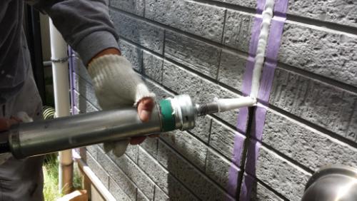 Person applies sealant to a brick wall using a caulking gun, with purple painter's tape masking the joint.
