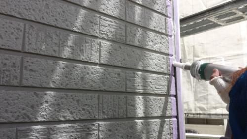 20130828-110929 | 外壁塗装は神奈川県横浜市の塗装職人 Person spraying white paint onto a brick exterior with a spray gun; purple painter’s tape lines the edge beside scaffolding in the background.