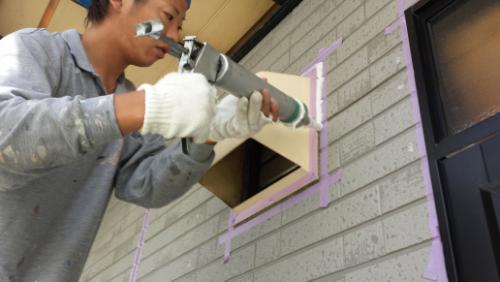 20130828-111207 | 外壁塗装は神奈川県横浜市の塗装職人 Person wearing gloves uses a power drill on a window opening, with purple painter's tape outlining the frame on a gray exterior wall.