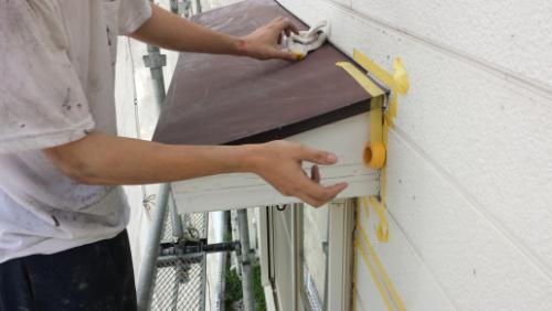 20130831-093258 | 外壁塗装は神奈川県横浜市の塗装職人 Construction worker attaching a brown metal panel to a white exterior wall, taping edges with yellow painter's tape while standing on scaffolding.