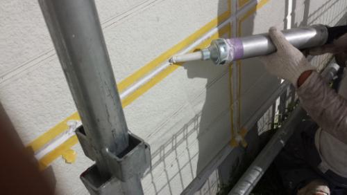Worker wearing gloves applies a yellow sealant/paint along an exterior wall using a caulking gun on scaffolding.