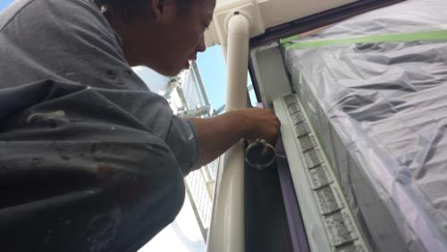 20130902-104732 | 外壁塗装は神奈川県横浜市の塗装職人 Worker in a gray shirt paints a window frame from the outside, with plastic sheeting masking the area around the window.