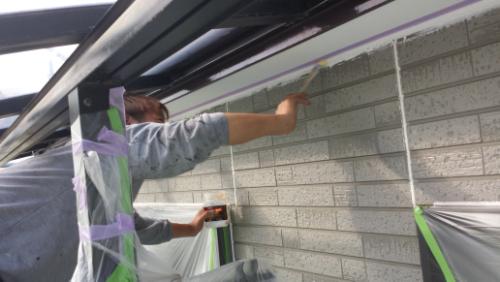 20130902-154606 | 外壁塗装は神奈川県横浜市の塗装職人 Person paints an exterior brick wall under a balcony, with masking tape and plastic sheeting protecting nearby surfaces.