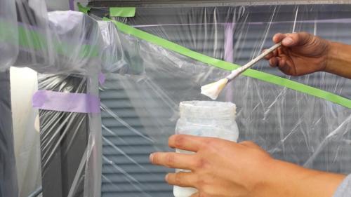 20130902-154830 | 外壁塗装は神奈川県横浜市の塗装職人 Hands brush white material onto a jar inside a plastic-covered workspace.