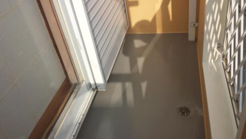 Narrow hallway or balcony with a metal shutter on the left, railing on the right, sunlit shadows on a gray floor, and a floor drain near the bottom right.