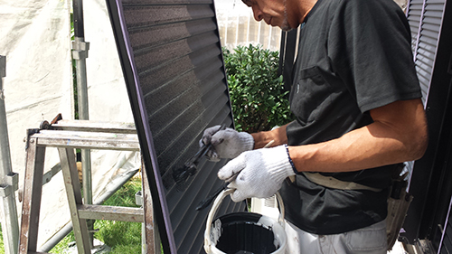koushin_07amado2 | 外壁塗装は神奈川県横浜市の塗装職人 Maintenance worker wearing gloves applies coating to a ribbed metal panel outdoors, with a bucket and ladder nearby.