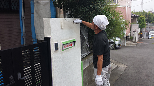 koushin_07monpei1 | 外壁塗装は神奈川県横浜市の塗装職人 Worker wearing gloves and a head covering paints a white textured exterior wall with a roller on a residential street street-side fence nearby.