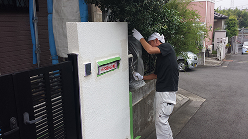 koushin_07monpei2 | 外壁塗装は神奈川県横浜市の塗装職人 Two workers repainting a white exterior wall panel near a gate, with masking tape and painting tools visible.