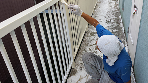 koushin_10fefensu | 外壁塗装は神奈川県横浜市の塗装職人 Person in a blue shirt and gray hoodie painting a white railing on a narrow balcony with a paintbrush.