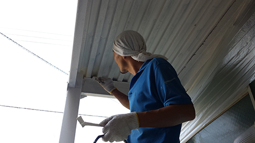 koushin_10fenakanuri | 外壁塗装は神奈川県横浜市の塗装職人 Worker in a blue shirt paints the underside of an outdoor overhang with a brush, wearing gloves and a head covering.