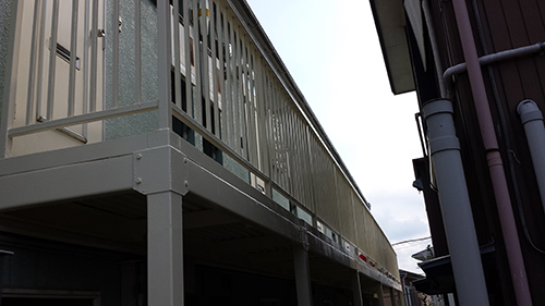 koushin_13fensu | 外壁塗装は神奈川県横浜市の塗装職人 Second-story balcony with light gray railing and supports, seen from below, attached to a house with dark siding on the right.