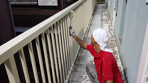Worker in a red shirt and white headscarf painting a metal railing with a roller on a narrow outdoor balcony.
