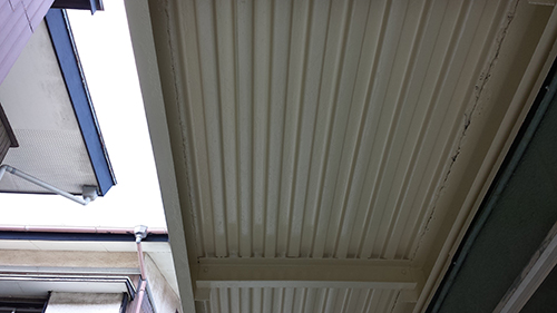 koushin_13urauwanuri | 外壁塗装は神奈川県横浜市の塗装職人 Low-angle view of a beige corrugated metal awning/ceiling between two buildings, with walls and windows visible nearby in an outdoor alley.