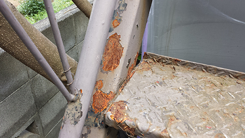 koushin_29hagare1 | 外壁塗装は神奈川県横浜市の塗装職人 Close-up of a rusted metal staircase railing and diamond-plate step with peeling orange rust, outdoors near a concrete block wall.
