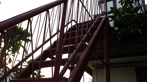 koushin_29sabidome | 外壁塗装は神奈川県横浜市の塗装職人 Red metal exterior staircase with diagonal supports and railing, climbing beside a building with some green leaves nearby