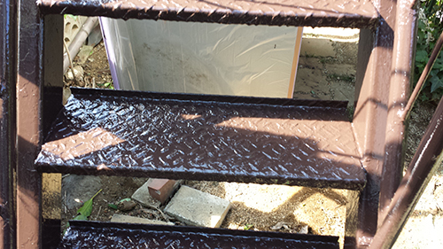 koushin_30tetsu4 | 外壁塗装は神奈川県横浜市の塗装職人 Close-up of a rusted brown metal staircase with diamond-plate steps outdoors, with bricks and a tarp in the background.