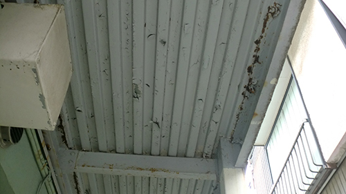 koushin_tosoumae2 | 外壁塗装は神奈川県横浜市の塗装職人 Low-angle view of a corrugated metal ceiling with rust stains and peeling paint in an indoor space near a windowed wall.