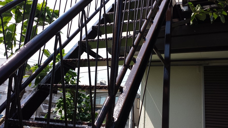 Outdoor black metal staircase with railing leading to an upper level, wet steps and surrounding greenery beside a building wall.