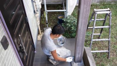 Man kneeling on a tiled porch, applying sealant to a wooden post while a ladder and bucket sit nearby