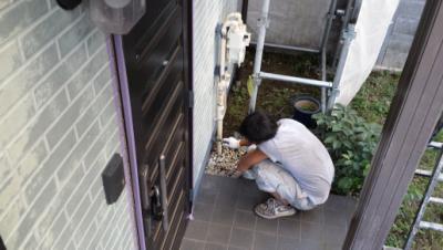 Person crouched on a tiled entryway outside a door, leaning toward a pipe or utility panel and working with their hands.